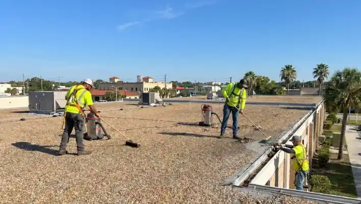 Wide-angle photograph of a professional three-person commercial roofing crew wearing full safety harnesses and high-visibility vests. Two workers are sweeping and vacuuming a gravel-ballasted flat roof, and a third worker is cleaning debris from a gutter on the edge, under a clear blue sky in Lakeland FL.